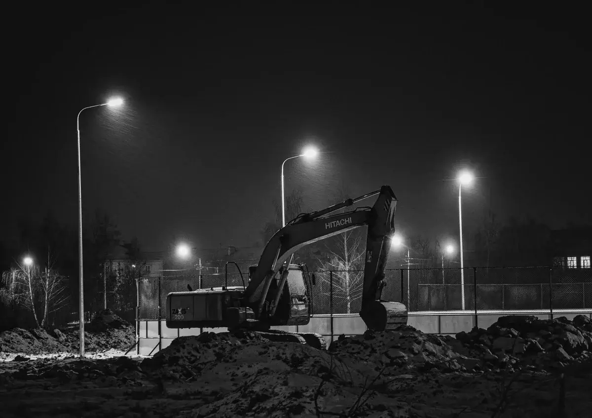 Black-and-white photo of an excavator at night under streetlights.