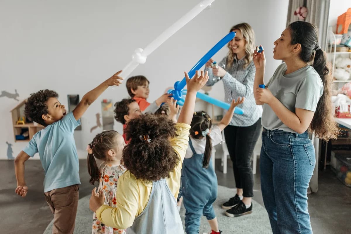 Children and teachers playing with balloons indoors in a kindergarten classroom.