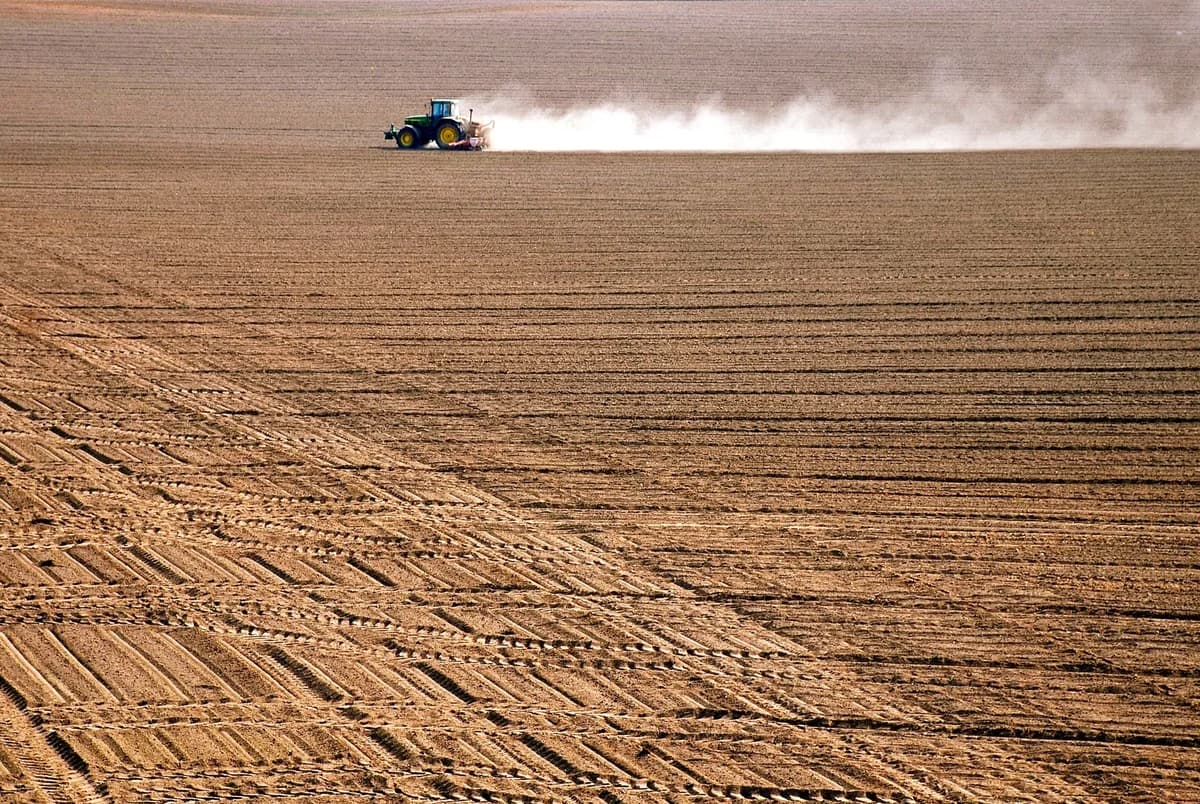 A tractor drives across a large ploughed field, kicking up dust.