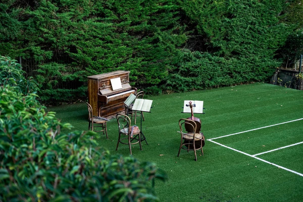 Old piano and chairs with music stands on green lawn near tennis court