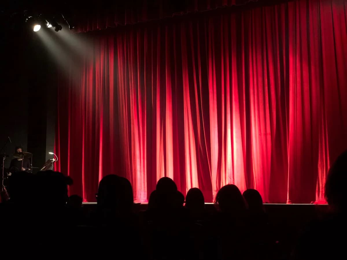 A theatre stage with red curtains lit by spotlights and audience silhouettes in front.
