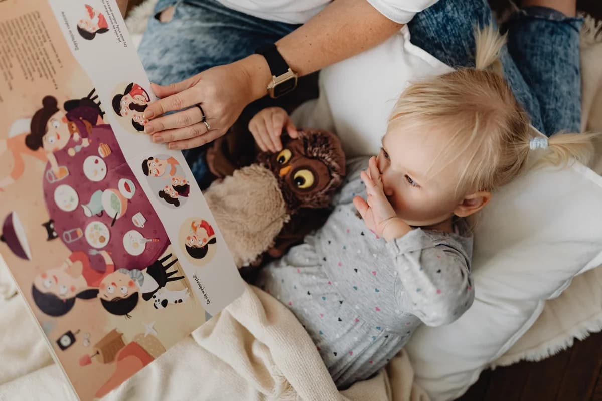 A child reading a picture book with a parent while holding an owl plush toy.