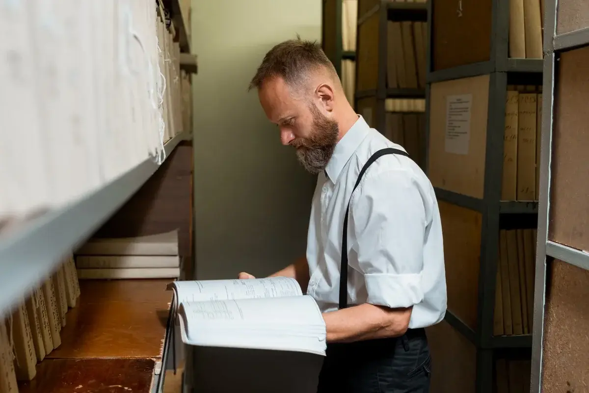 A bearded man in suspenders looking through a large book in an archive room.