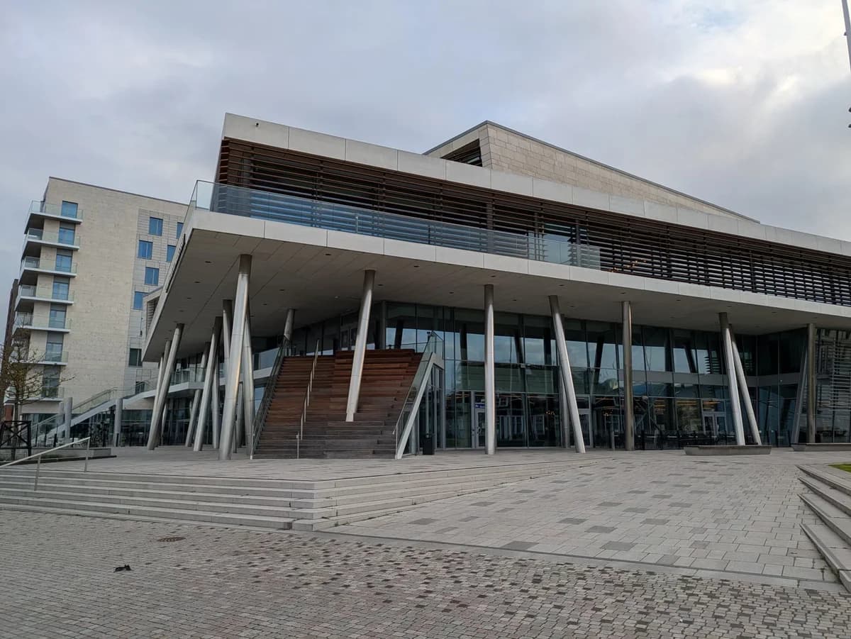A modern glass-fronted building with a wide staircase entrance and overhanging roof.