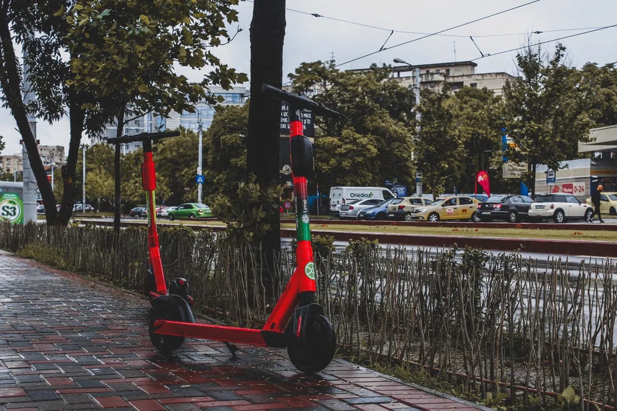 A red electric scooter parked on a wet sidewalk beside a city street.