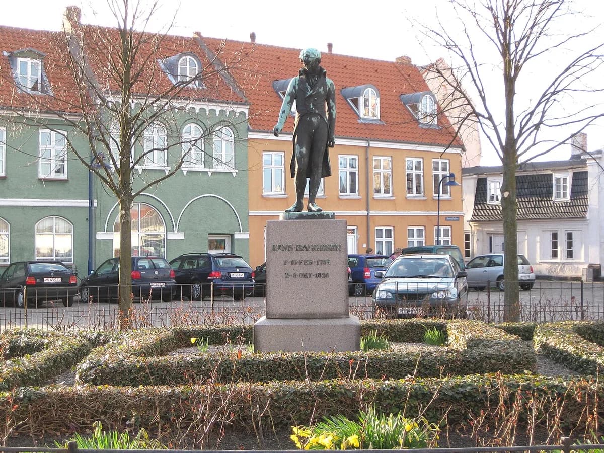 Statue of Jens Baggesen in a small garden with Danish buildings and parked cars behind it.