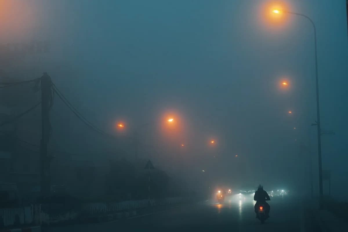 A motorcyclist rides down a foggy street at night under glowing streetlights.