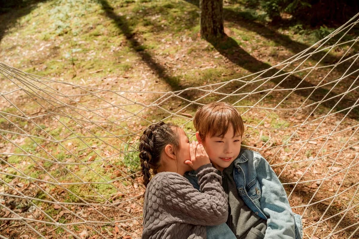 Two children sit in a hammock, with one whispering to the other in a sunny outdoor setting.